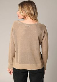 Beige trui met een ontspannen pasvorm, geribbeld zoom en raglanmouwen. Kenmerkt zich door een klein zilveren knoopdetail aan de achterkant van de halslijn.