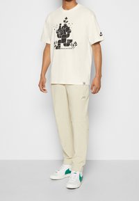 Krémové bavlněné tričko s černým grafickým designem, v kombinaci se světle béžovými tepláky a bílými teniskami s zelenými akcenty.