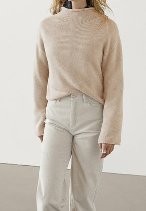Femme portant un pull en maille beige par-dessus une chemise sombre, associé à un pantalon en velours côtelé clair taille haute, debout devant un fond blanc.