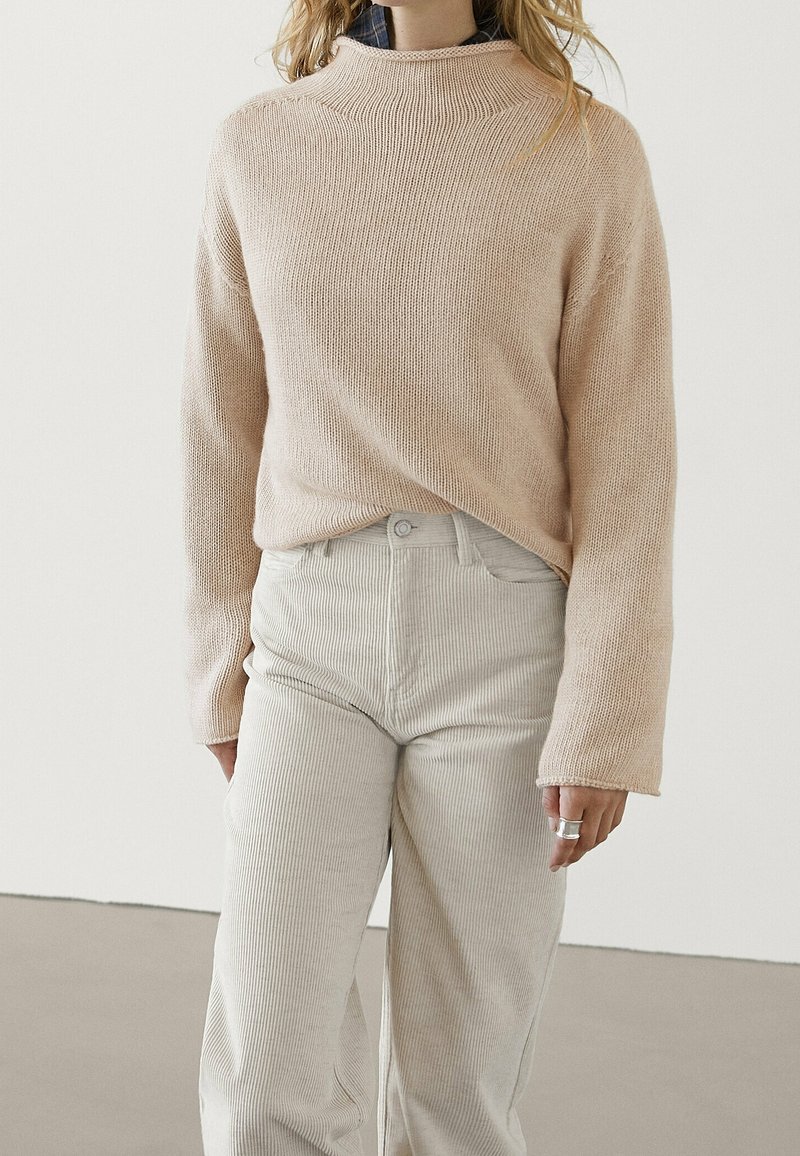 Femme portant un pull en maille beige par-dessus une chemise sombre, associé à un pantalon en velours côtelé clair taille haute, debout devant un fond blanc.