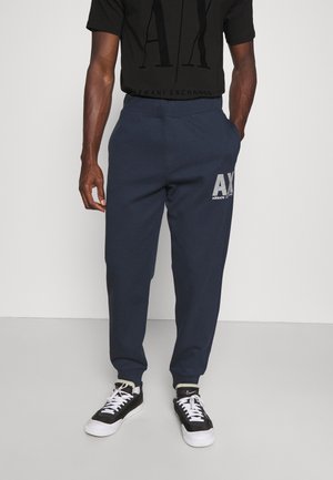 Mann trägt dunkelblaue Armani Exchange Jogginghose, schwarzes Armani Exchange T-Shirt und schwarz-weiße Nike Sneakers, steht vor weißem Hintergrund.