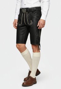 Pantalones cortos de cuero negro con costuras amarillas y acentos bordados, combinados con calcetines de color crema hasta la rodilla y zapatos marrones con cordones.