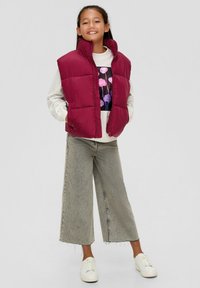 Bordeauxfarbener Puffer-Weste mit Reißverschluss und hohem Kragen über einem hellgrauen Pullover, kombiniert mit weit geschnittenen, verblassten Jeans und weißen Sneakern.