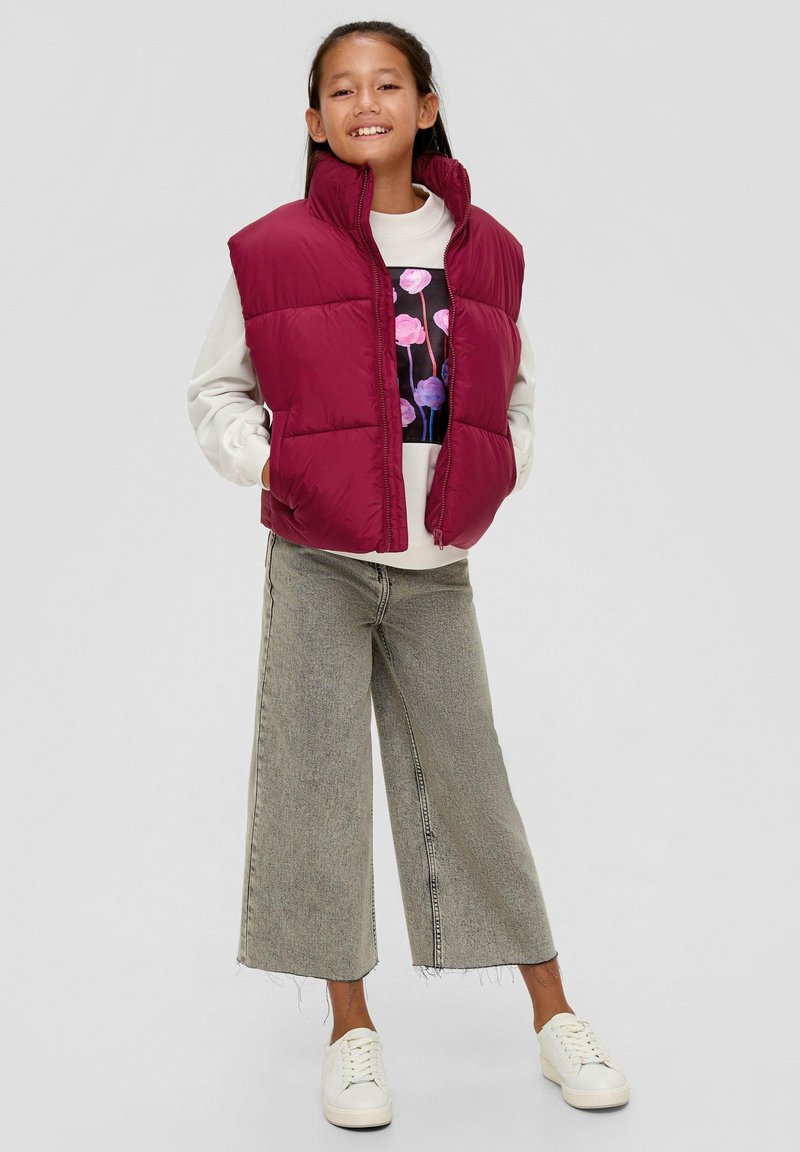 Bordeauxfarbener Puffer-Weste mit Reißverschluss und hohem Kragen über einem hellgrauen Pullover, kombiniert mit weit geschnittenen, verblassten Jeans und weißen Sneakern.