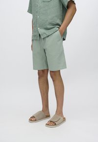 Light green linen shirt with short sleeves and pocket, paired with matching textured shorts and beige slide sandals with a padded strap.