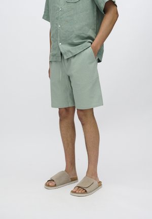 Light green linen shirt with short sleeves and pocket, paired with matching textured shorts and beige slide sandals with a padded strap.