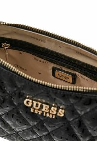 Sac à main noir matelassé avec un logo et une fermeture éclair dorés. L'intérieur présente une doublure claire avec un motif répétitif "GUESS" et une petite poche zippée.