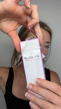 Vrouw pakt een Mugler Starlicious parfumdoosje uit met een doorzichtige plasticfolie tegen een effen achtergrond.