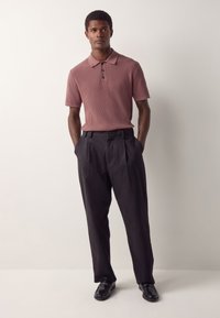 Korte mouwen, geribbelde polo in stofroze met een drukknoopsluiting met drie knopen, gecombineerd met geplooide zwarte broek en zwarte loafers.