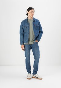 Veste en denim bleu foncé avec un motif texturé, accompagnée d'un t-shirt vert clair, d'un jean bleu et de baskets blanches avec des accents noirs.