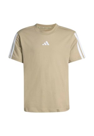 Ragazzo biondo che indossa una t-shirt beige Adidas e pantaloncini con strisce bianche e logo, in piedi davanti a uno sfondo chiaro e uniforme.