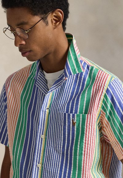Ριγέ πουκάμισο με κοντά μανίκια που διαθέτει κάθετες ρίγες σε πορτοκαλί, πράσινο, μπλε και κόκκινο χρώμα. Περιλαμβάνει τσέπη στο στήθος και λεπτομέρειες με κουμπιά.