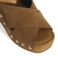 Les sandales en daim marron présentent des courroies croisées, une semelle en bois, et des clous dorés le long des bords, avec des détails cousus sur les courroies.