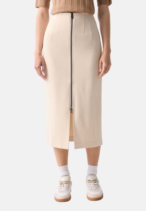 Femme portant une jupe midi beige avec une fermeture éclair devant, des chaussettes blanches, des baskets blanches et un haut côtelé marron.