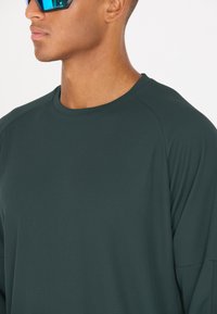 Dunkelgrünes, strukturiertes Langarmshirt mit rundem Ausschnitt. Verfügt über einen atmungsaktiven Stoff und eine lockere Passform. Reflektierende Sonnenbrille wird getragen.