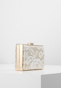 Kleine rechthoekige gouden clutch met witte bloemrijke kantlaag, rechtopstaand op een witte ondergrond tegen een effen achtergrond.