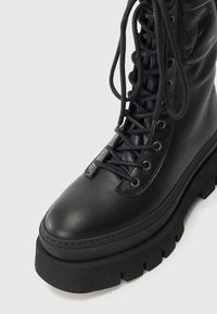 Schwarze Lederkampfstiefel mit dicker Gummisohle, runder Fußspitze, Schnürsystem und strukturierten Akzenten entlang des Schafts.