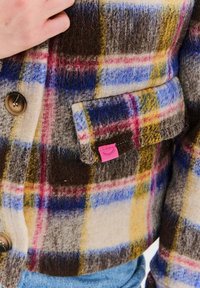 Vlněná kostkovaná bunda s hnědou, modrou, žlutou a červenou barvou. Výrazné zapínání na knoflíky a růžová textilní visačka s ikonou řas.