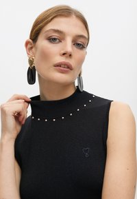 Top negro sin mangas con cuello alto, que cuenta con tachuelas en color oro rosa alrededor del cuello y un pequeño detalle de corazón bordado. Tela suave y texturada.