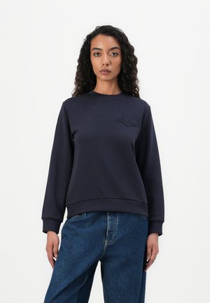 Marineblå hettegenser med rund hals, ribbestrikkede mansjetter og kant, samt en teksturert logousmykning på brystet. Kombinert med vide blå denimjeans.