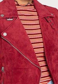 Červená semišová motocyklová bunda se stříbrnými detaily, diagonálním zipem a vnitřním pruhovaným topem v oranžové, červené a černé barvě, který má texturovaný povrch.