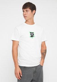 Hvit bomulls t-skjorte med et svart og grønt grafisk logo på brystet. Korte ermer og en klassisk rund hals design.