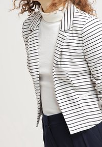 Blazer à rayures blanc et bleu marine, avec un col cranté, une coupe ajustée et un tissu doux, porté avec un col roulé blanc en dessous.