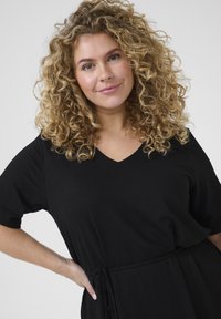 Black dress with short sleeves, V-neckline, textured fabric, and a drawstring waist. Model presents the dress with hands on hips.