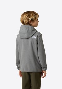 Šedá fleecová mikina s kapucňou, s bielym logom na zadnom ramene. Hladká textúra a voľný strih, doplnená olivovo zelenými nohavicami.