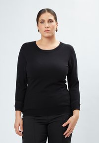 Schwarzer Strickpullover mit runder Ausschnitt und Dreiviertelärmeln, ausgestattet mit strukturierten Bündchen und einer taillierten Passform. Kombiniert mit schwarzen Hosen.