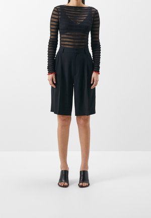 Vrouw die een zwarte doorzichtige, gestreepte top met lange mouwen, zwarte knielange short met hoge taille en zwarte open-teen mule hakken draagt.
