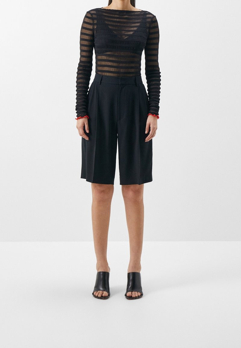 Femme portant un haut noir transparent à manches longues rayé, un short noir taille haute longueur genou et des mules à talons noires avec orteils ouverts.