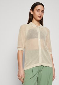 Lys beige mesh cardigan med krage, korte ærmer, knaplukning og tekstureret strikkemønster. Båret over grønne bukser.