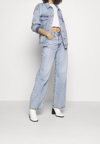 Světlě modrá džínová bunda, bílý crop top, světlé modré džíny s širokými nohavicemi a bílé kotníkové boty na podpatku na neutrálním pozadí.