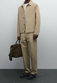 Giacca in tela beige con bottoni neri, pantaloni beige abbinati e scarpe nere. Una borsa a viaggio in pelle marrone è tenuta in una mano.