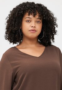 Femme aux cheveux bouclés et aux boucles d'oreilles créoles, portant un haut brun décolleté en V. Elle regarde le spectateur avec confiance sur un fond clair.