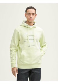 Sudadera verde claro con cordón, bolsillo frontal y texto de logo blanco. Mangas largas y puños acanalados. Combinada con jeans de denim oscuro.