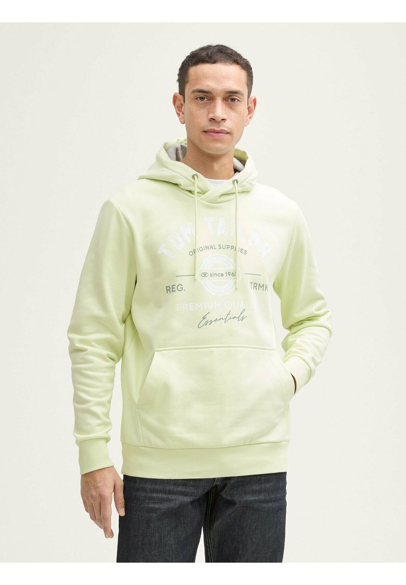 Sudadera verde claro con cordón, bolsillo frontal y texto de logo blanco. Mangas largas y puños acanalados. Combinada con jeans de denim oscuro.