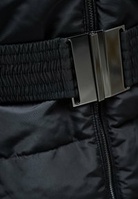 Detalle de chaqueta acolchada negra con un broche negro brillante y tela recogida alrededor de la cintura. La cremallera delimita el frente, añadiendo textura.