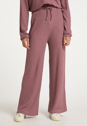 Vrouw die mauve geribbelde wijde broek met een trekkoortaille en witte sneakers draagt, staand tegen een effen achtergrond.