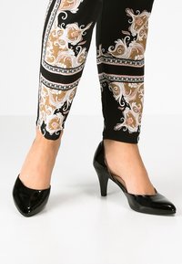 Escarpins en cuir verni noir avec un bout pointu et un talon modeste, associés à un legging noir et doré à motifs représentant des designs floraux.