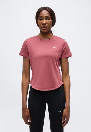 Kobieta stoi zwrócona przodem, ubrana w różowy sportowy t-shirt z krótkim rękawem i czarne legginsy, oba z białymi logo Nike.