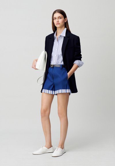Blazer marine sur une chemise à carreaux bleus et blancs, associé à un short bleu avec un ourlet rayé. Chaussures blanches et sac à main blanc.
