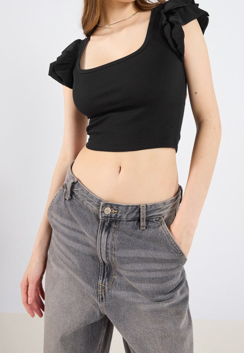 Femme portant un haut court noir à manches volantées et un jean gris ample taille haute, une main dans la poche, sur fond uni.