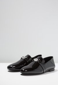 Chaussures en cuir verni noir avec un bout pointu, une semelle plate et une boucle décorative sur le dessus. Texture lisse et design élégant.