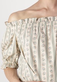 Blouse dénudée sur les épaules en tissu crème léger avec des motifs floraux en bleu et orange, dotée d'un col froncé élastiqué et de manches bouffantes.
