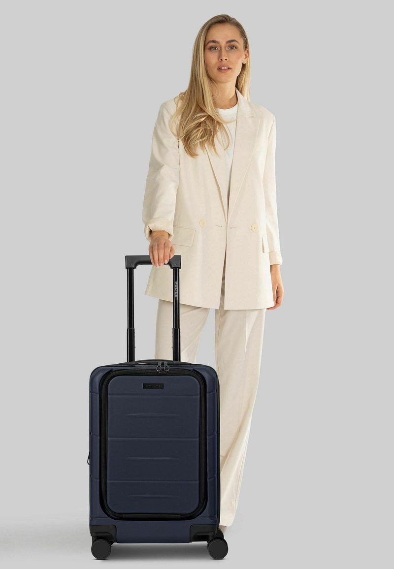 Donna in completo beige che tiene la maniglia di una valigia trolley blu navy con manico telescopico esteso su sfondo grigio.