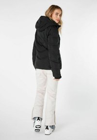 Giacca a vento nera con cappuccio, caratterizzata da una texture liscia e tasche con zip. Abbinata a pantaloni da sci di colore chiaro e scarponi da sci con dettagli distintivi.