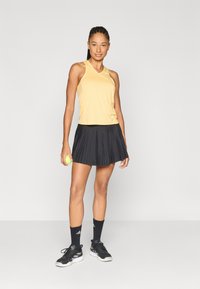 Mouwloos geel sporttopje met een losse pasvorm, gecombineerd met een geplooide zwarte rok. Model houdt een groene tennisbal vast en draagt zwarte schoenen en sokken.