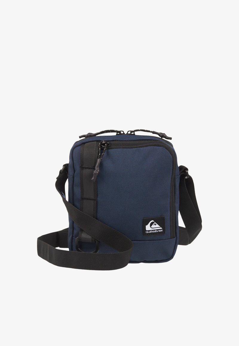 Petit sac bandoulière bleu marine avec sangle noire réglable, compartiment principal zippé et écusson logo noir à l'avant.